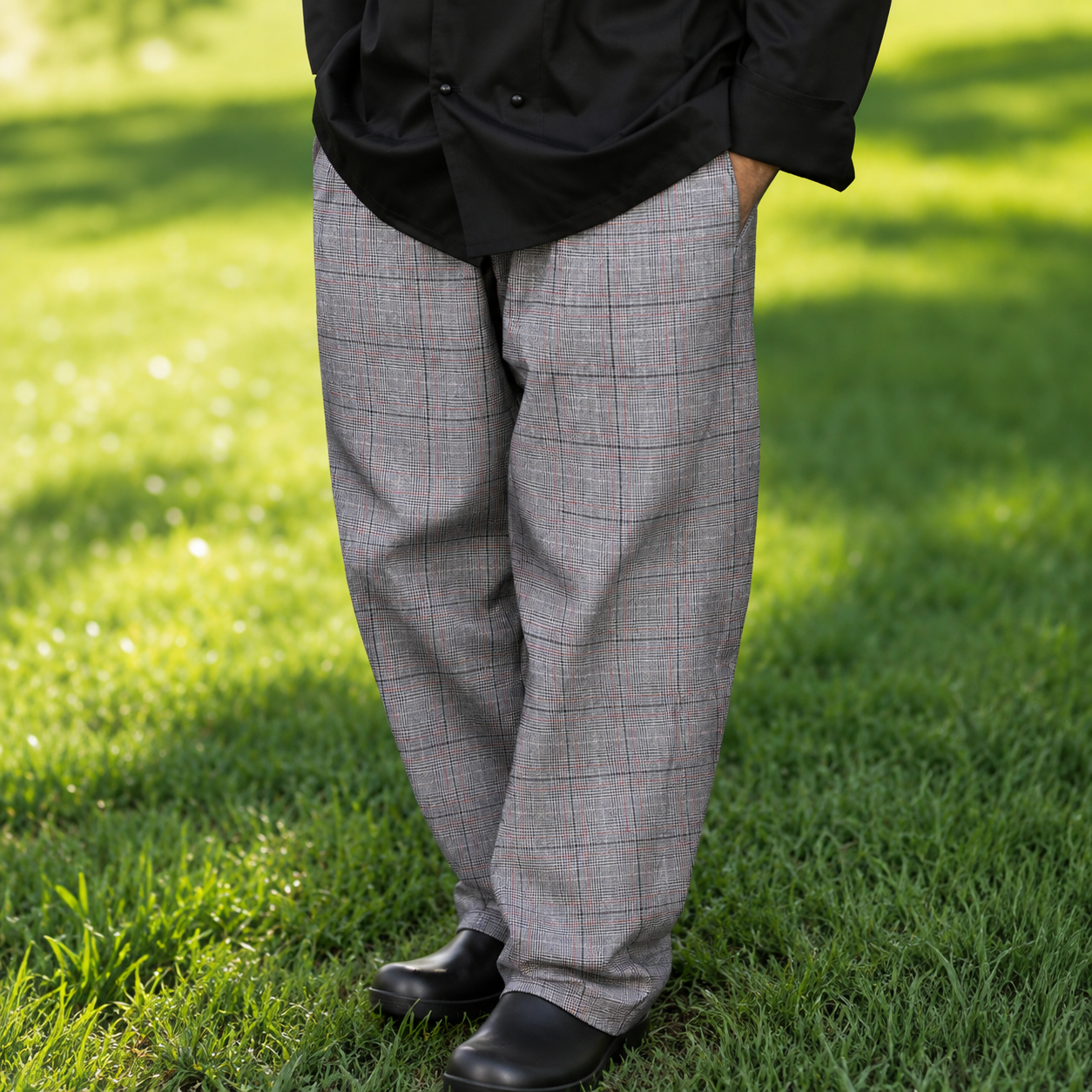 A person standing on green grass wearing gray windowpane plaid trousers, a black shirt, and black shoes.