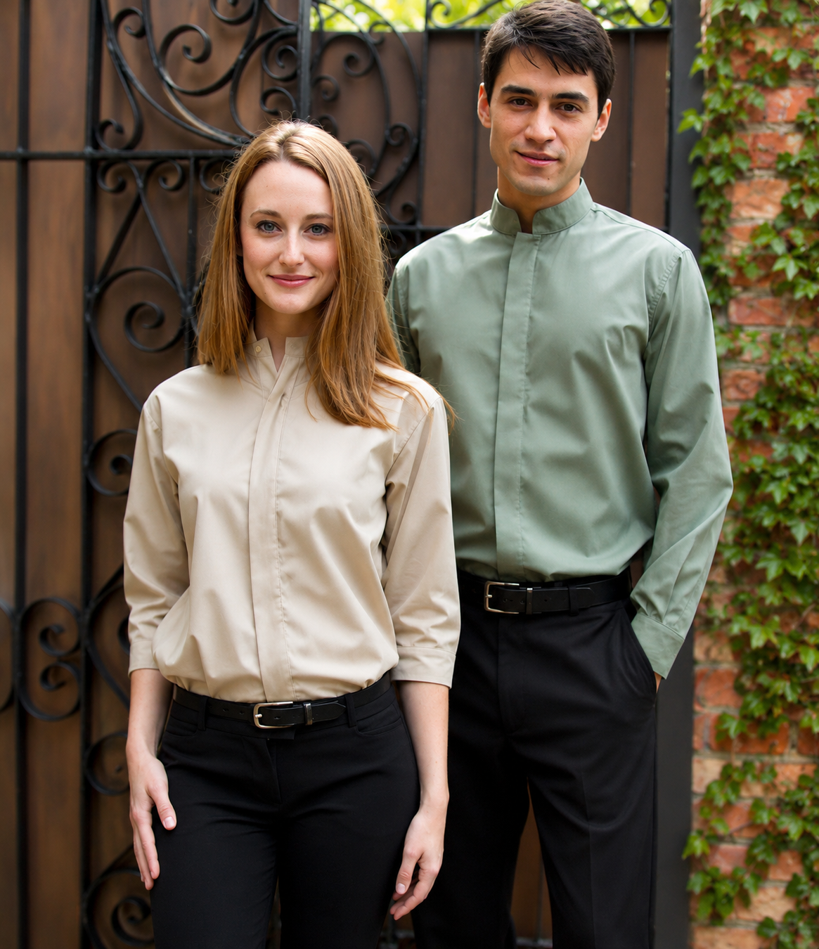 A man and a woman standing outdoors in front of a decorative black metal gate and brick wall with ivy. The woman wears a light beige long-sleeve button-up shirt with a high collar and black pants, while the man wears a sage green long-sleeve button-up shirt with a high collar and black pants.