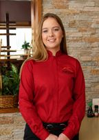 A woman wearing a red long-sleeve button-up chef jacket with a black belt and black pants, standing in a restaurant setting with a stone wall background.