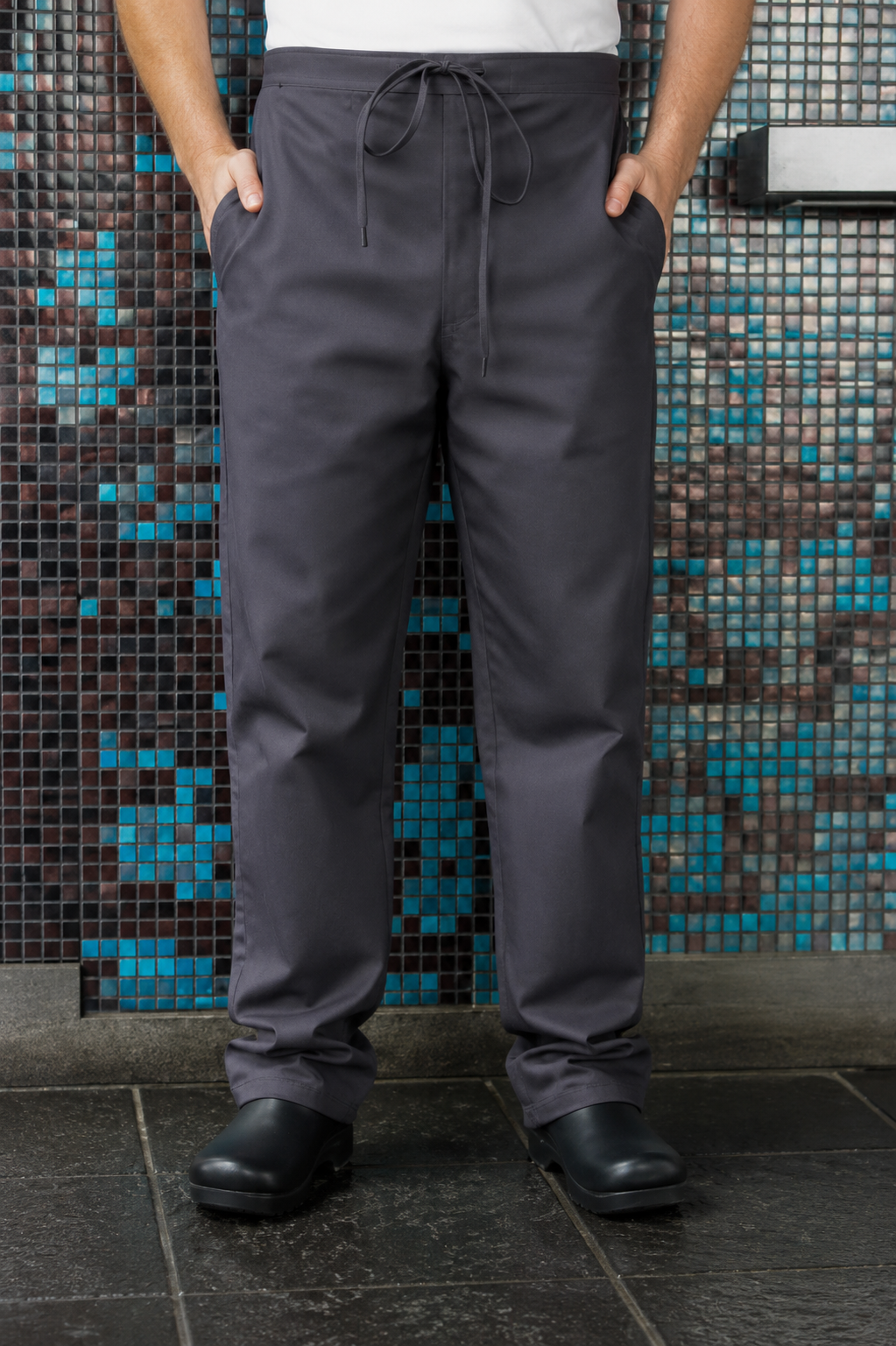 A person wearing dark gray drawstring scrub pants with side pockets, paired with black clogs, standing in front of a blue and black mosaic tile wall.